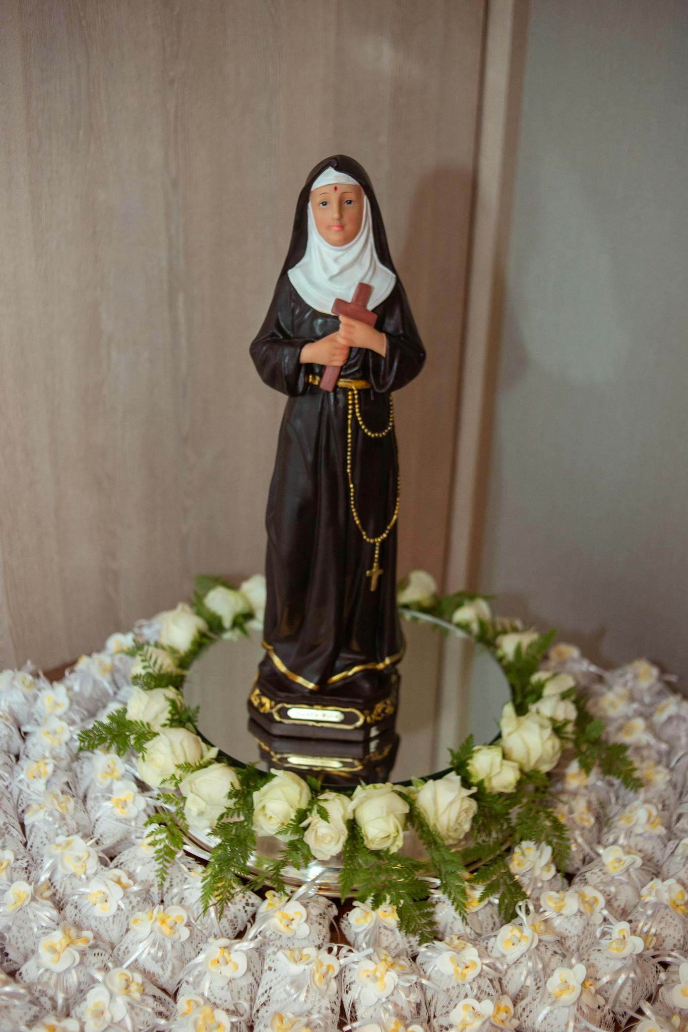 A nun figurine on a mirror, encircled by white flowers and rosary beads, symbolizing devotion.