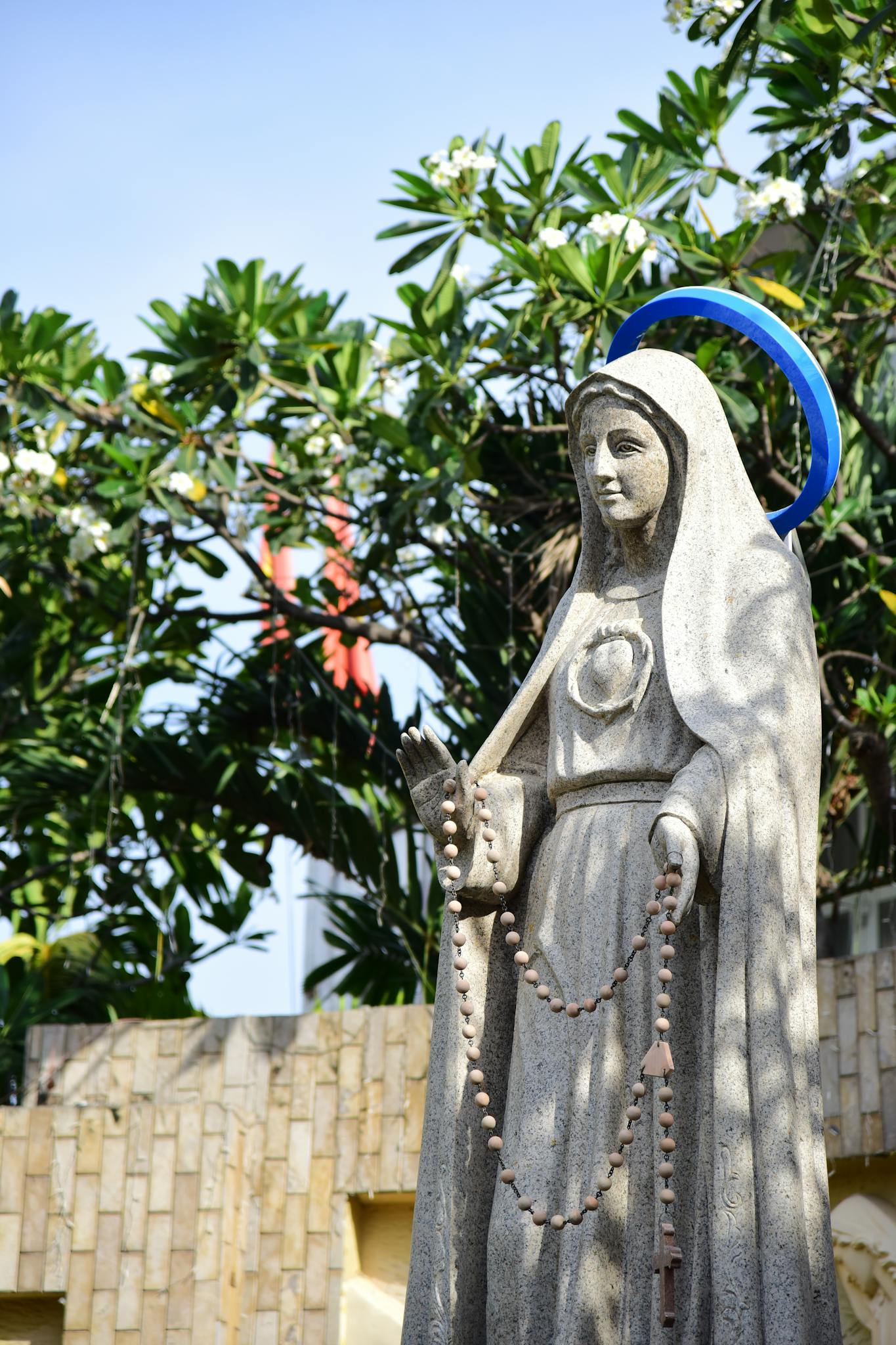 A stone statue of a religious figure holding a rosary in a garden setting with trees.